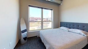 Habitación de hotel con un ventanal grande, una cama con cabecero gris y una tabla de surf apoyada en la pared.
