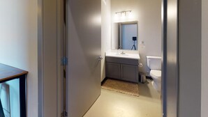 Un couloir étroit mène à une salle de bain équipée d’un lavabo blanc, d’un miroir et de toilettes.