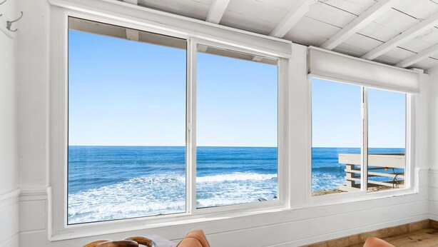 A room with large windows overlooking the ocean, wooden flooring, and a view of a beach and a building across the water. - Single Family Beachfront Home with a Spa P2031-0 (Oceanside)