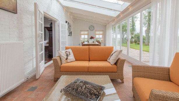 A bright, open-plan living area with a sofa, a coffee table, and a view of the outdoors through large windows. - ‘Prachtig gelegen vrijstaand landhuis met grote zonnige tuin’ (Hansweert)