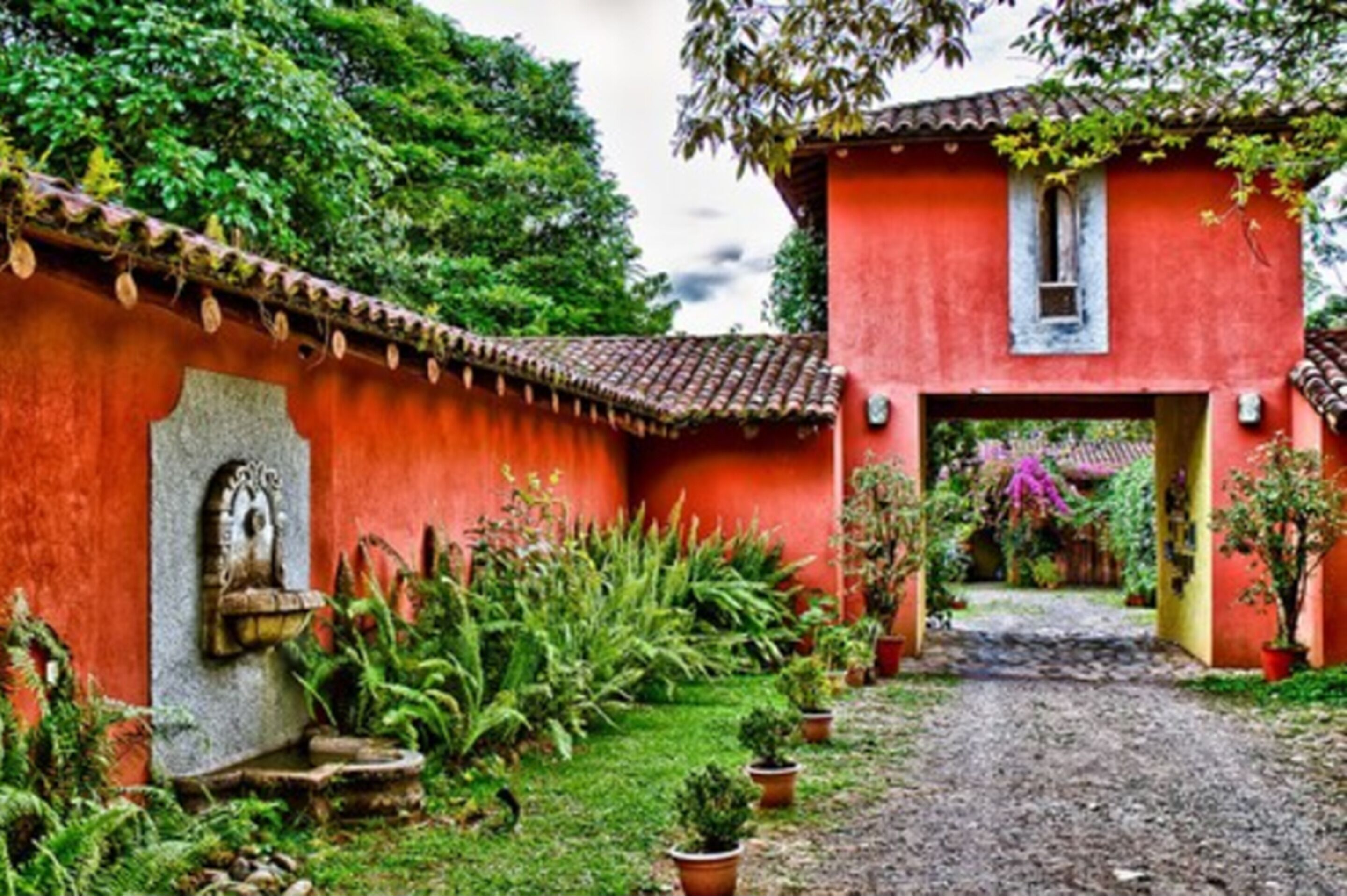 A red building with a tiled roof, a stone wall with a niche, and a gravel path leading to a door.