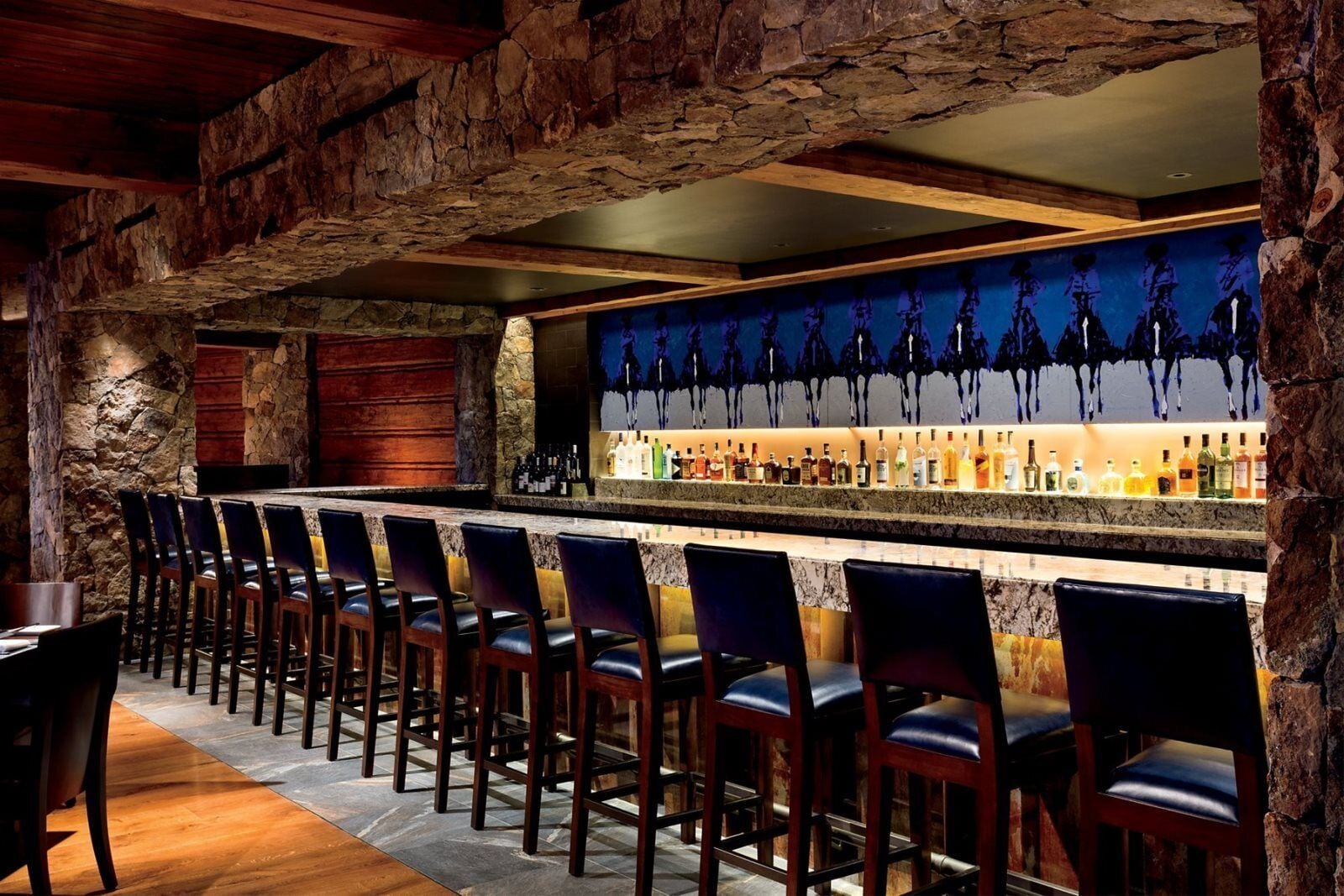 A bar area with stone walls, a long counter with bar stools, and a wall-mounted liquor display.