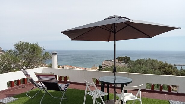 Una terraza con dos sillas blancas, una mesa y un paraguas grande, con vista al mar.