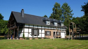 Un bâtiment traditionnel à colombages, doté d’un toit en pente et d’une cheminée, entouré d’une pelouse verdoyante et d’arbres.