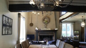 Un coin repas avec une longue table, des chaises en osier, une cheminée ornée d’une plaque portant l’inscription « CAMPAINE » et un lustre.