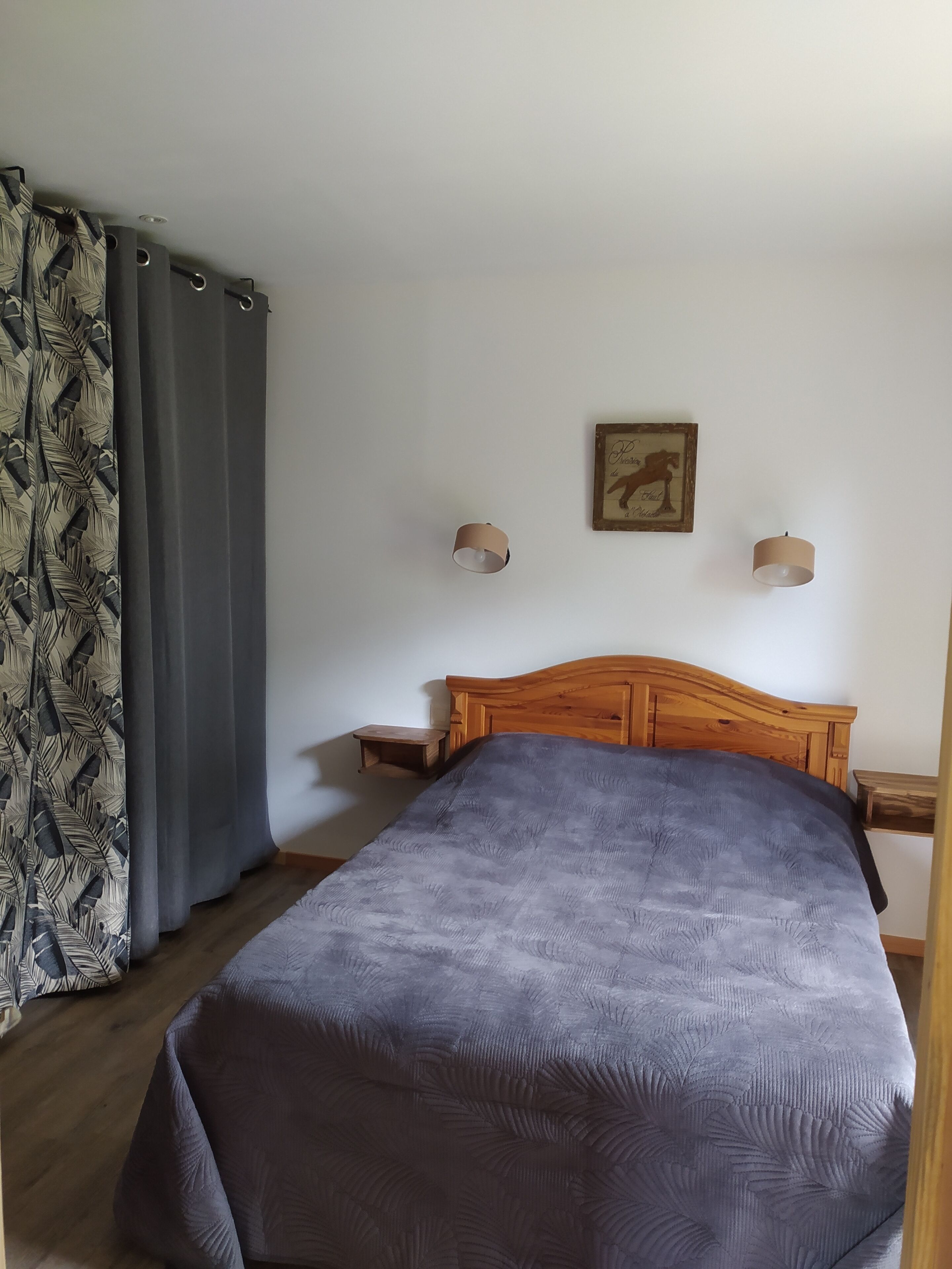 A bedroom with a wooden bed, grey curtains, a small bedside table, and a wall-mounted picture.