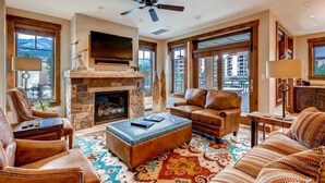 A spacious living room with a stone fireplace, leather sofas, and a large window offering a city view. - Spacious 4 Bedroom Vacation Rental in Convenient Location with Scenic Views, Pool, Hot Tub (Breckenridge)