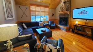 A cozy living room with a stone fireplace, wooden ceiling, and a television displaying 'Live Lodge Life'. - LIVE LODGE LIFE- Upscale decor, sleeps 15, hot tub, bikes, 10x SHARC, amazing!! (Sunriver)