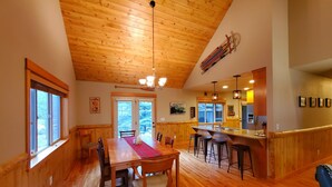 A spacious dining area with a wooden table and chairs, a kitchen with a bar, and a view of the outdoors through large windows. - LIVE LODGE LIFE- Upscale decor, sleeps 15, hot tub, bikes, 10x SHARC, amazing!! (Sunriver)