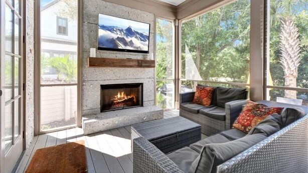 A sunlit patio with a stone fireplace, outdoor seating, and a TV mounted on a stone wall. - Ocean Oaks 19 - gorgeous home only a short walk to pool and beach (Jekyll Island)