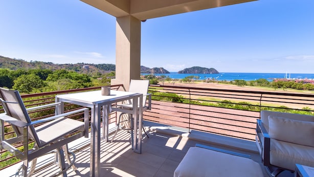 A balcony with a table and chairs, overlooking a coastal landscape with hills and the sea. - Los Suenos Resort Miramar 5B (Jacó)