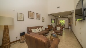A spacious living room with a brown leather sofa set, a glass coffee table, and a floor lamp. There are framed artworks on the walls and a television mounted on the wall. - Los Suenos Resort Veranda 1C (Jacó)