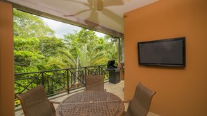 A balcony with a round table and chairs, a television mounted on the wall, and a grill in the background. - Los Suenos Resort Veranda 1C (Jacó)