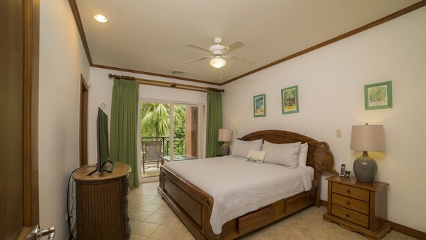 A bedroom with a large bed, a nightstand, a lamp, a ceiling fan, and a view of the outdoors. - Los Suenos Resort Veranda 1C (Jacó)