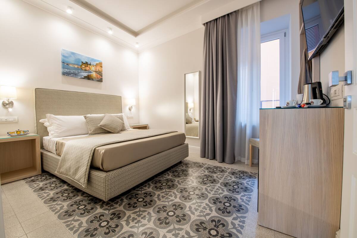 A hotel room with a bed, a nightstand, a flat-screen TV mounted on the wall, and a window with curtains.
