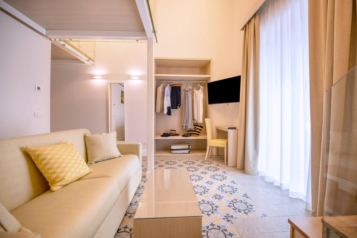 A modern hotel room with a sofa, a flat-screen TV, a small table, a wardrobe, and a chair.