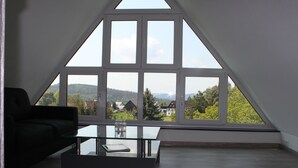 Un salon moderne avec une table basse en verre, un canapé noir et une grande fenêtre donnant sur un paysage boisé.