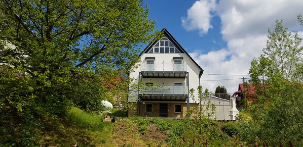 Set amidst lush greenery, this two-story home features a gabled roof and a balcony, blending modern design with a natural setting.