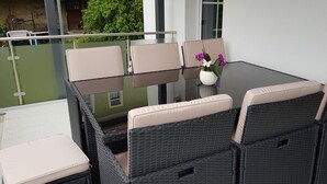 Une table à plateau en verre, quatre chaises beiges avec des coussins, un vase avec des fleurs violettes, et une vue sur un paysage verdoyant et un bâtiment en arrière-plan.