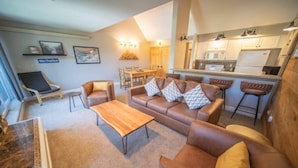 Un salon avec un canapé en cuir marron, une table basse en bois et un coin repas comprenant une table et des chaises. Il y a également une cuisine avec des meubles blancs et un réfrigérateur.