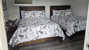 Une chambre avec deux lits en bois, chacun recouvert d’une courtepointe à motifs représentant des élans et des arbres. On y trouve deux lampes fixées au mur, une fenêtre avec des stores et un tableau encadré accroché au mur.