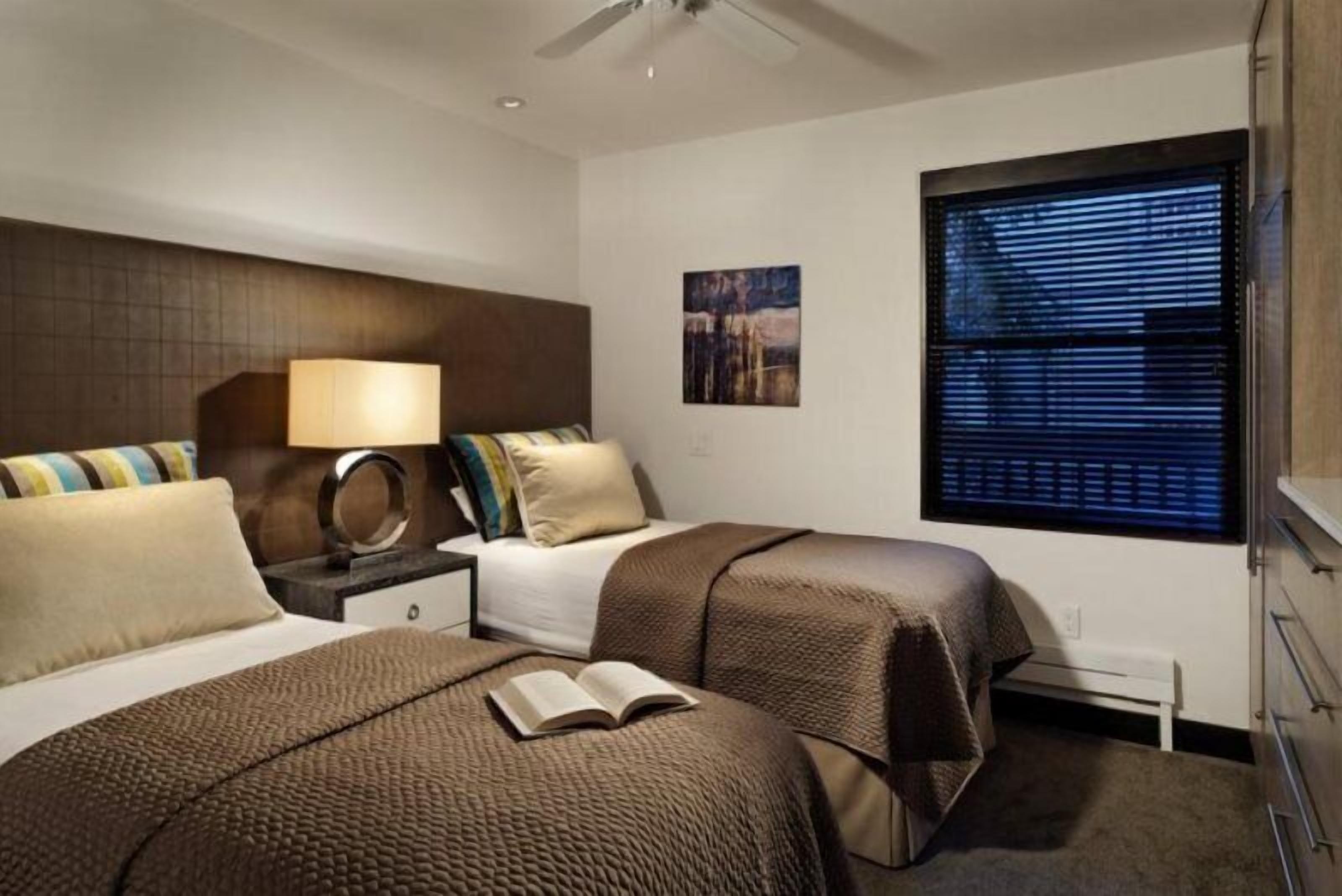 A hotel room with two beds, a nightstand, a lamp, a window with blinds, and a ceiling fan.