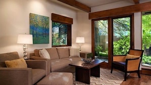 A modern living room with a sectional sofa, a coffee table, and a chair. There are two windows with a view of greenery outside. - Luxury 1 Bedroom Downtown Aspen Vacation Rental With Access to a Heated Pool, Hot Tubs, Game Room and Spa (Aspen)