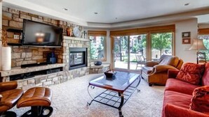A living room with a stone fireplace, a flat-screen TV, a glass coffee table, leather sofas, and large windows offering a view of the outdoors. - Ski in, Ski out 2 Bedroom Vacation Rental in the Heart of Lionshead Village (Vail)