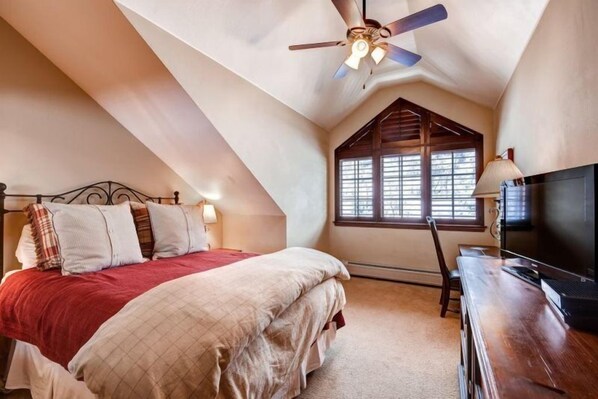 A bedroom with a sloped ceiling, a bed with a red blanket, a desk with a computer, and a window with blinds.