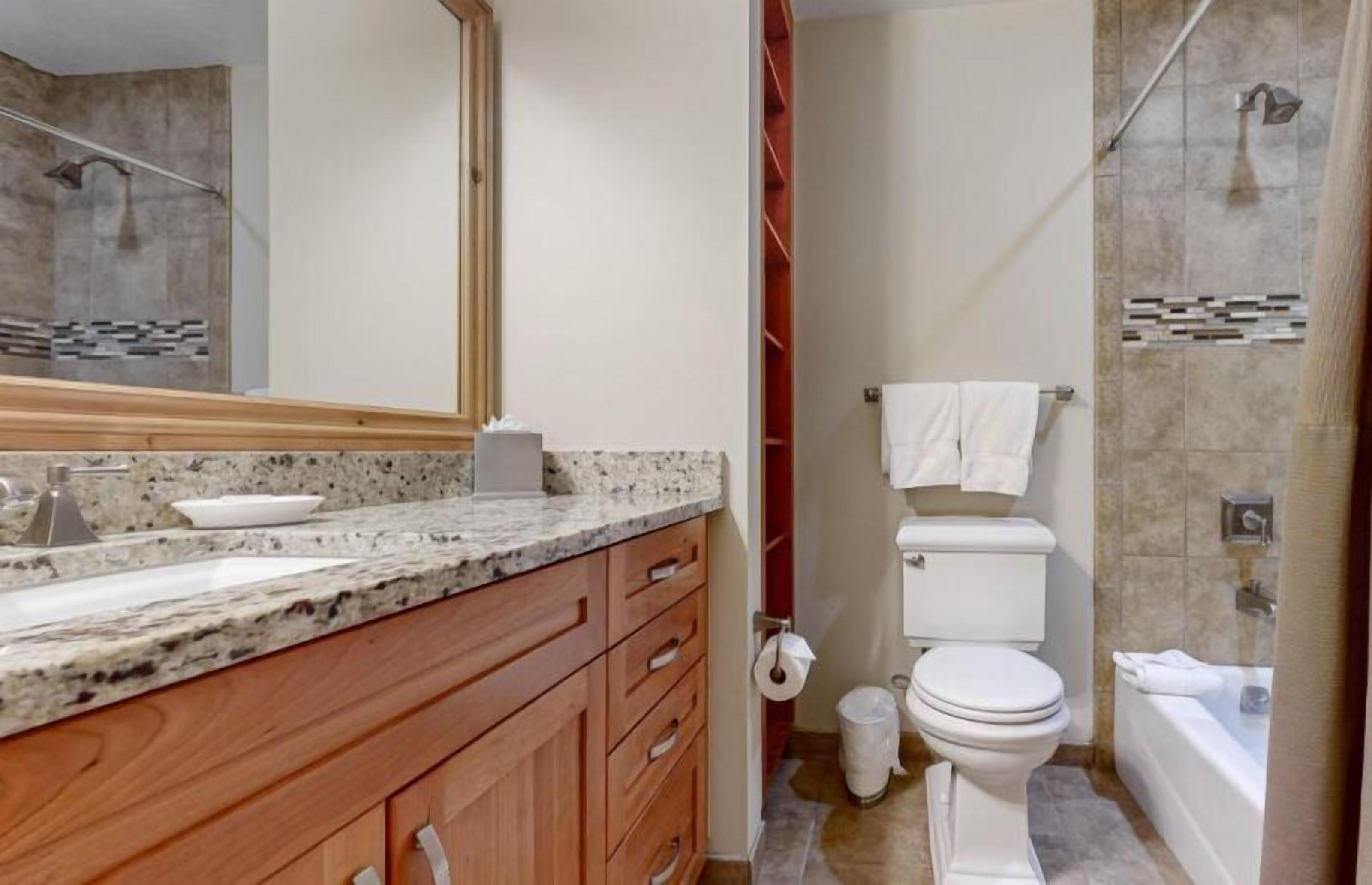 Une salle de bain avec un comptoir en marbre, un meuble-lavabo en bois, des toilettes blanches et une baignoire.