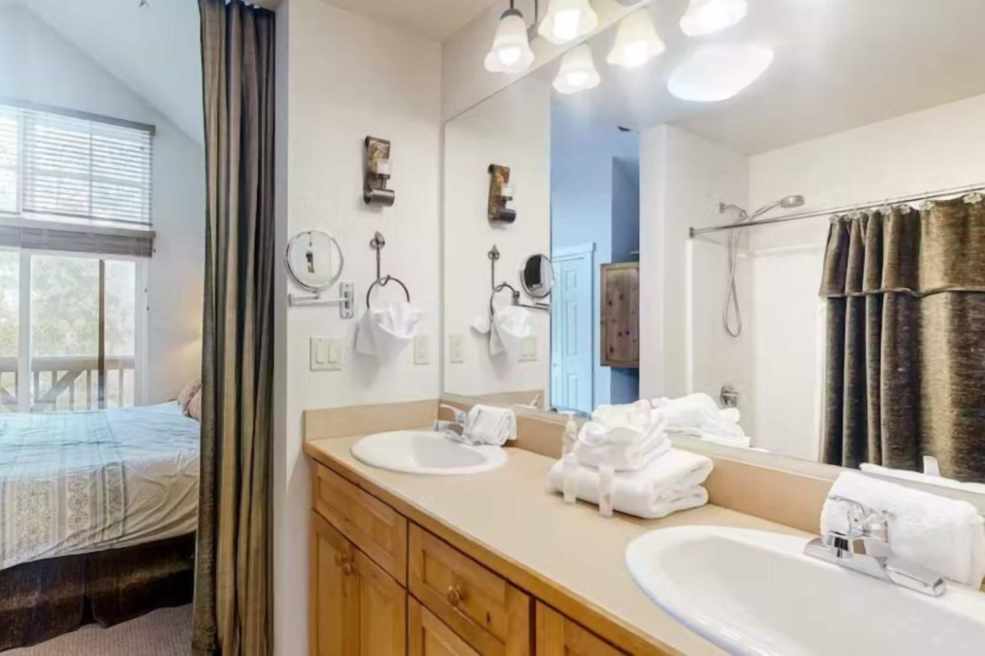 A hotel bathroom with a large mirror, two sinks, a shower, and a bed visible through a window.
