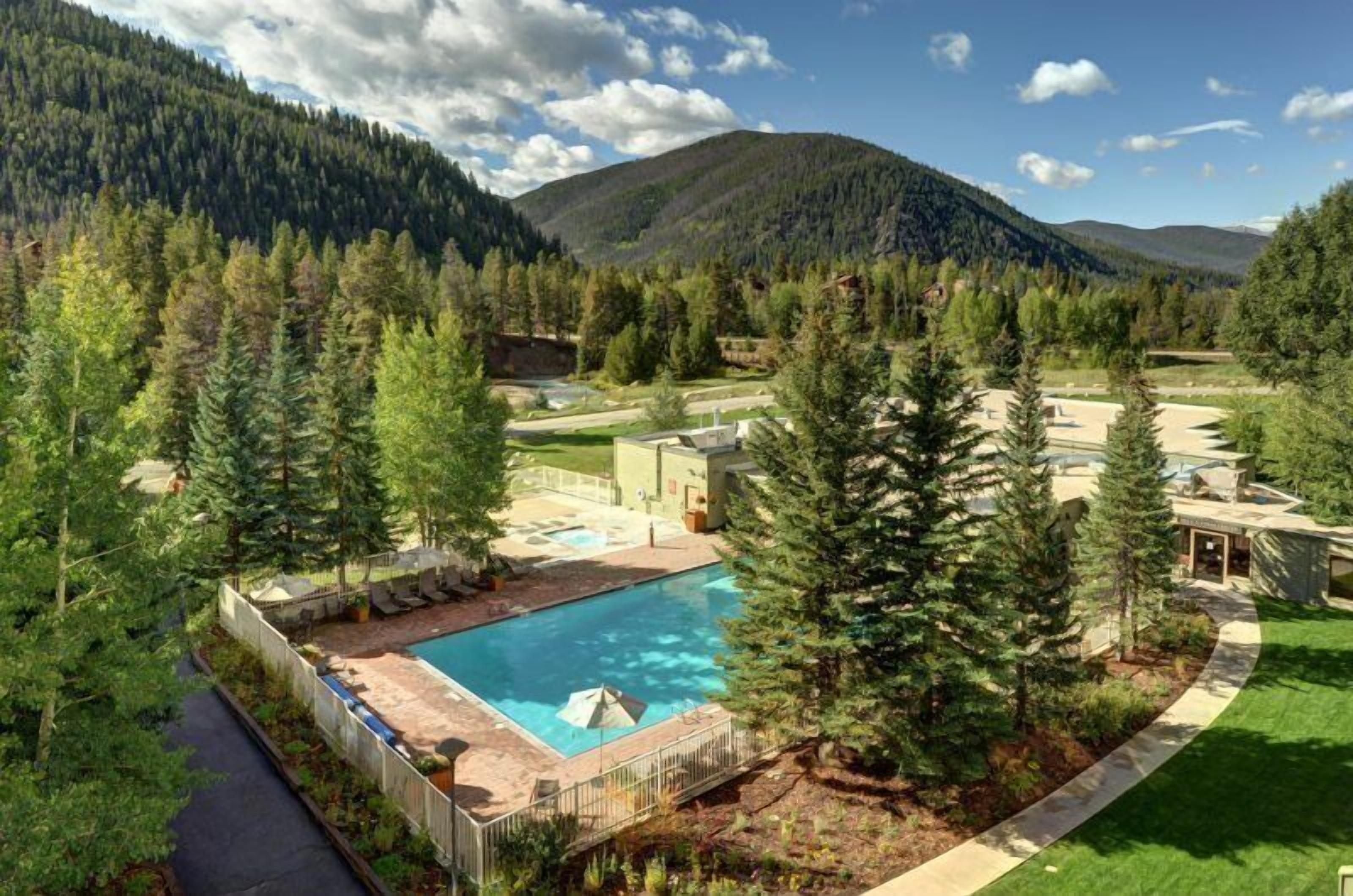 A residential area with a swimming pool surrounded by trees, a paved walkway, and a view of mountains.