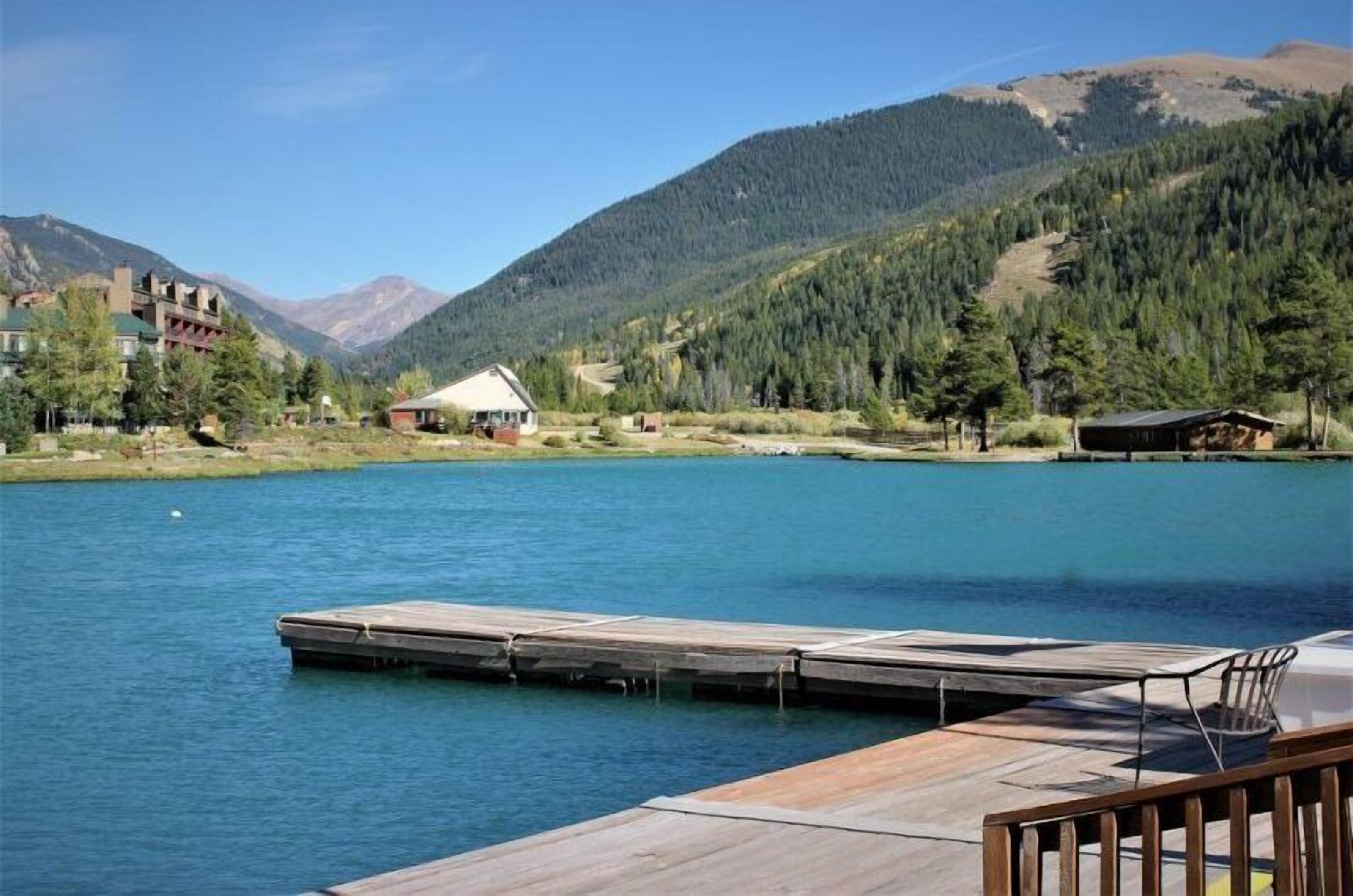 A lakeside view with a wooden dock, a small cabin, and a building with a green roof.