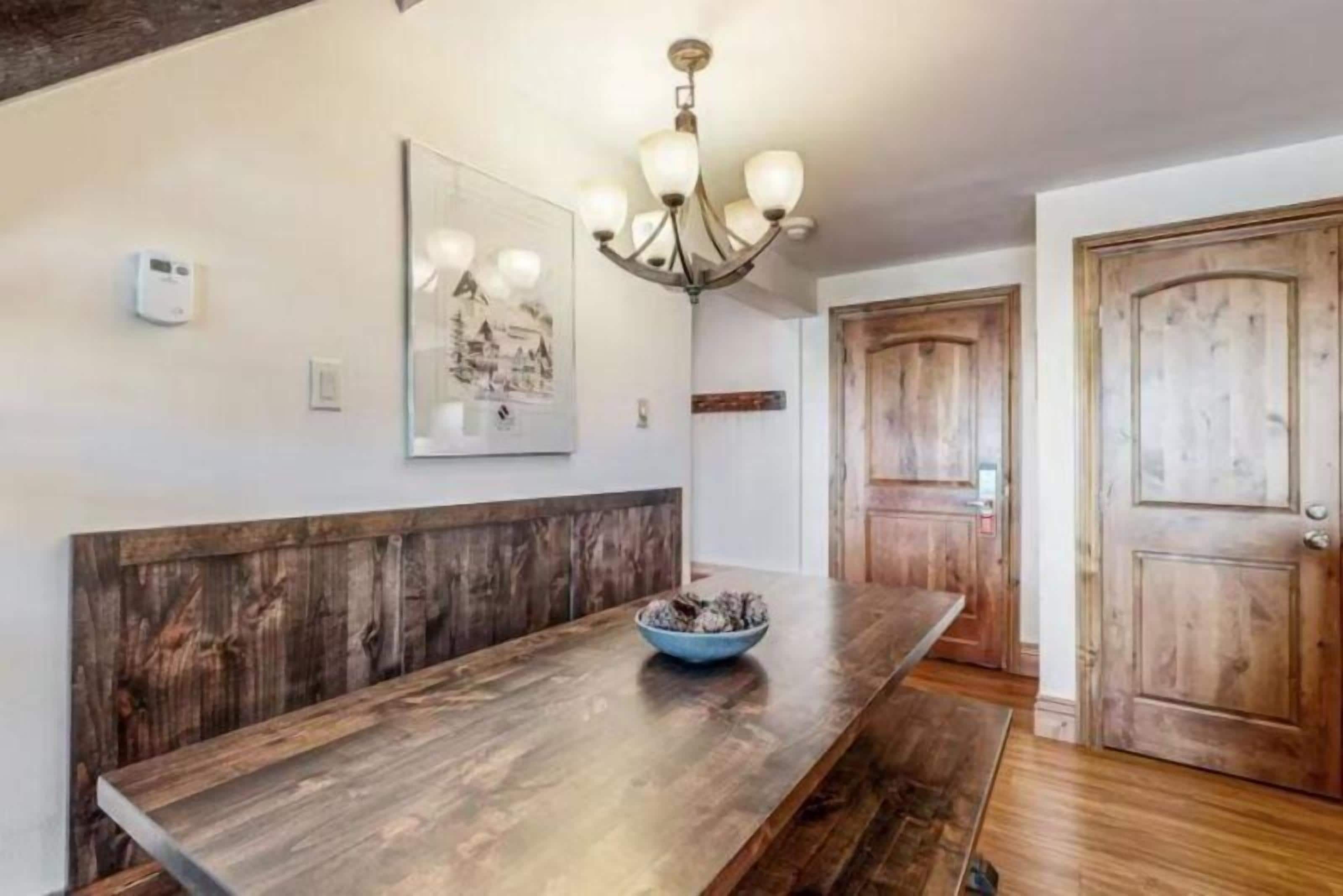 A wooden bench with a bowl, a chandelier, and a door in a room with white walls and a wooden floor.