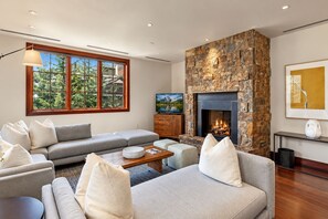 Un salon moderne avec une cheminée en pierre, une grande fenêtre donnant sur la forêt et un canapé gris.