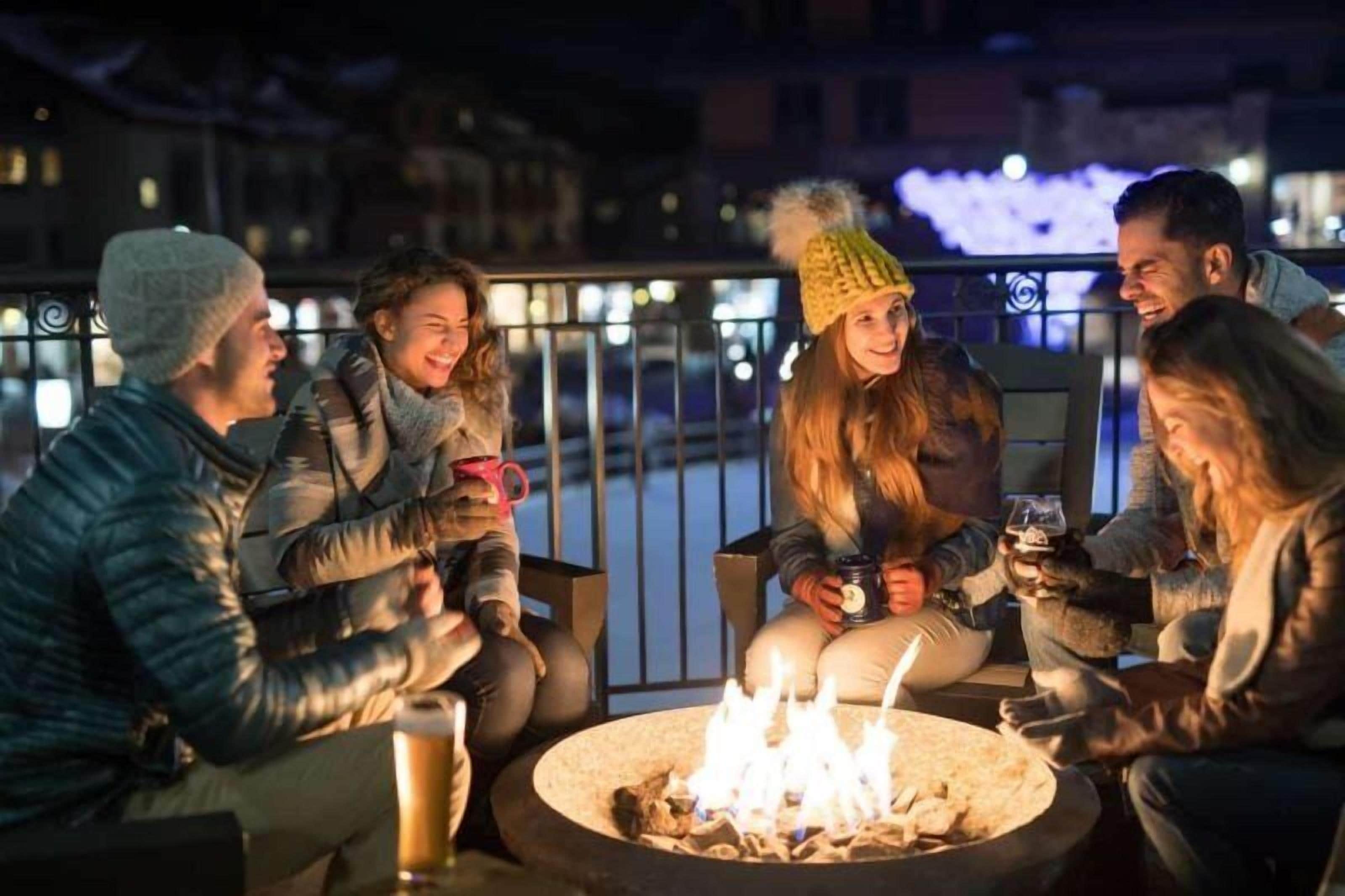 Un grupo de personas sentadas alrededor de un brasero por la noche, al aire libre, con una barandilla y edificios al fondo.