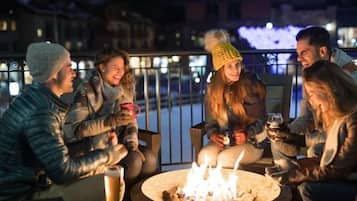 Un grupo de personas sentadas alrededor de un brasero por la noche, al aire libre, con una barandilla y edificios al fondo.