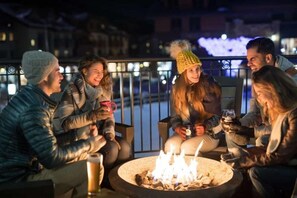 A group of people sitting around a fire pit at night, outdoors, with a railing and buildings in the background.