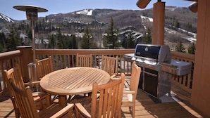 Eine Holzterrasse mit rundem Tisch und Stühlen, einem Grill und Blick auf eine Berglandschaft.