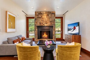 A living room with a stone fireplace, a sofa, two yellow armchairs, a coffee table with flowers, and a TV displaying a skier.