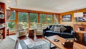 A spacious living room with a large window offering a view of a forest, a dark blue sofa, two armchairs, a coffee table, and a wooden sideboard with a lamp. - Lakeside Village 4 Bedroom Condo (Keystone)