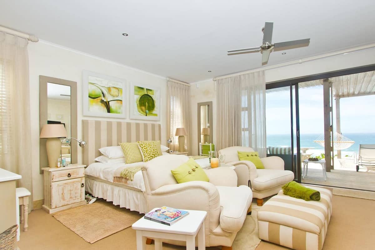 A spacious bedroom with a large bed, a sofa, a small table, and a view of the ocean through large sliding doors.