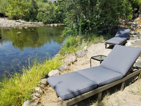 A serene outdoor seating area with two lounge chairs and a small table by a clear water body, surrounded by rocks and greenery. - Relax Swim and BBQ at Sierra River House (Three rivers)
