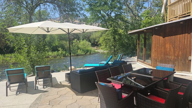 An outdoor seating area with wicker furniture, a table, and a large umbrella, overlooking a river and surrounded by greenery. - Relax Swim and BBQ at Sierra River House (Three rivers)