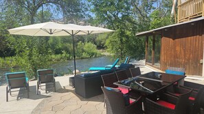 An outdoor seating area with wicker furniture, a table, and a large umbrella, overlooking a river and surrounded by greenery.