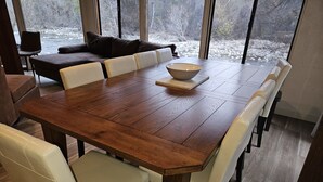 A large wooden dining table with white cushioned chairs, a bowl on the table, and a view of a river through large windows. - Relax Swim and BBQ at Sierra River House (Three rivers)