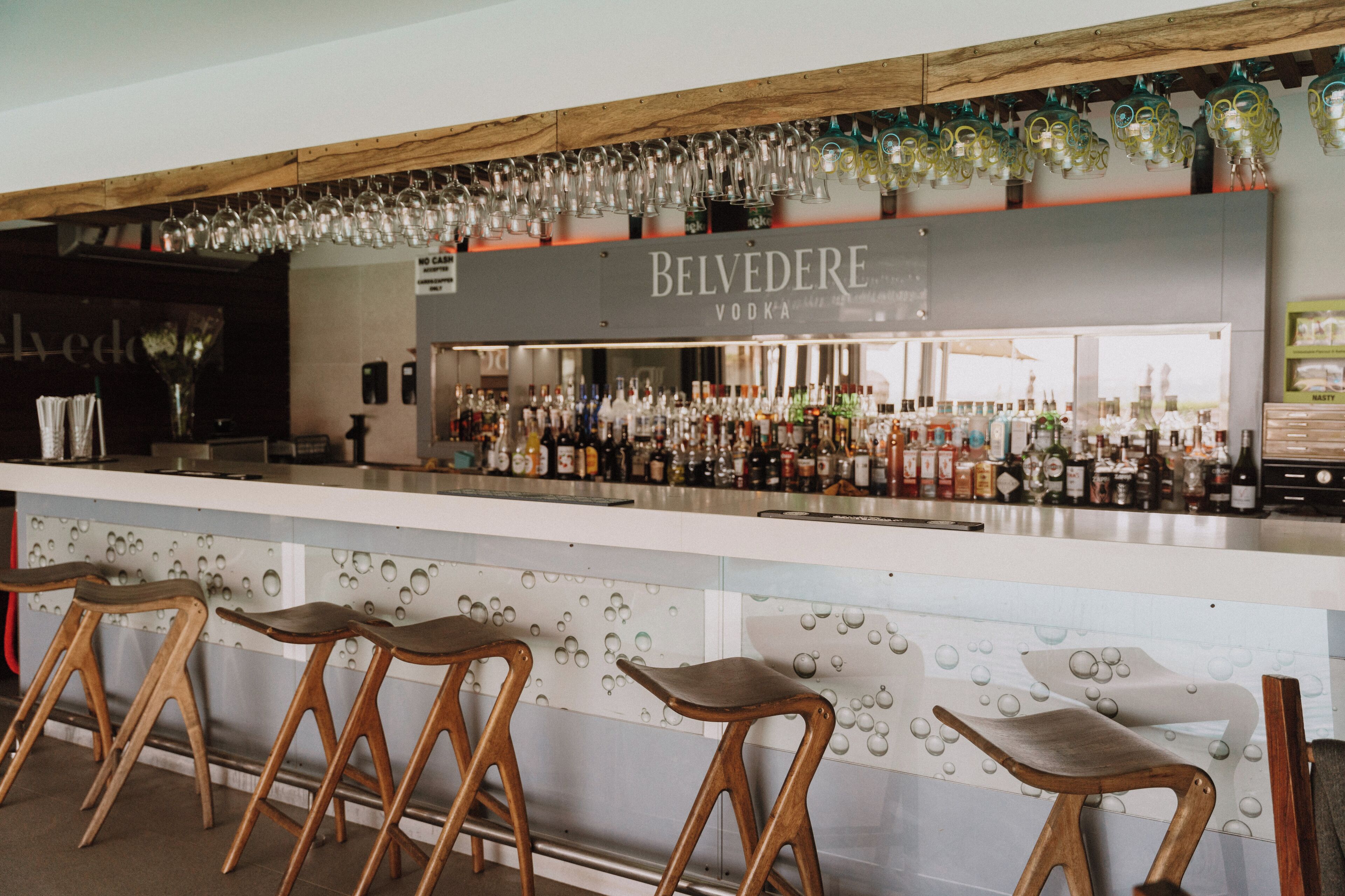 Uma área de bar com um balcão exibindo diversos frascos, uma placa para Belvedere Vodka e banquetas de madeira.