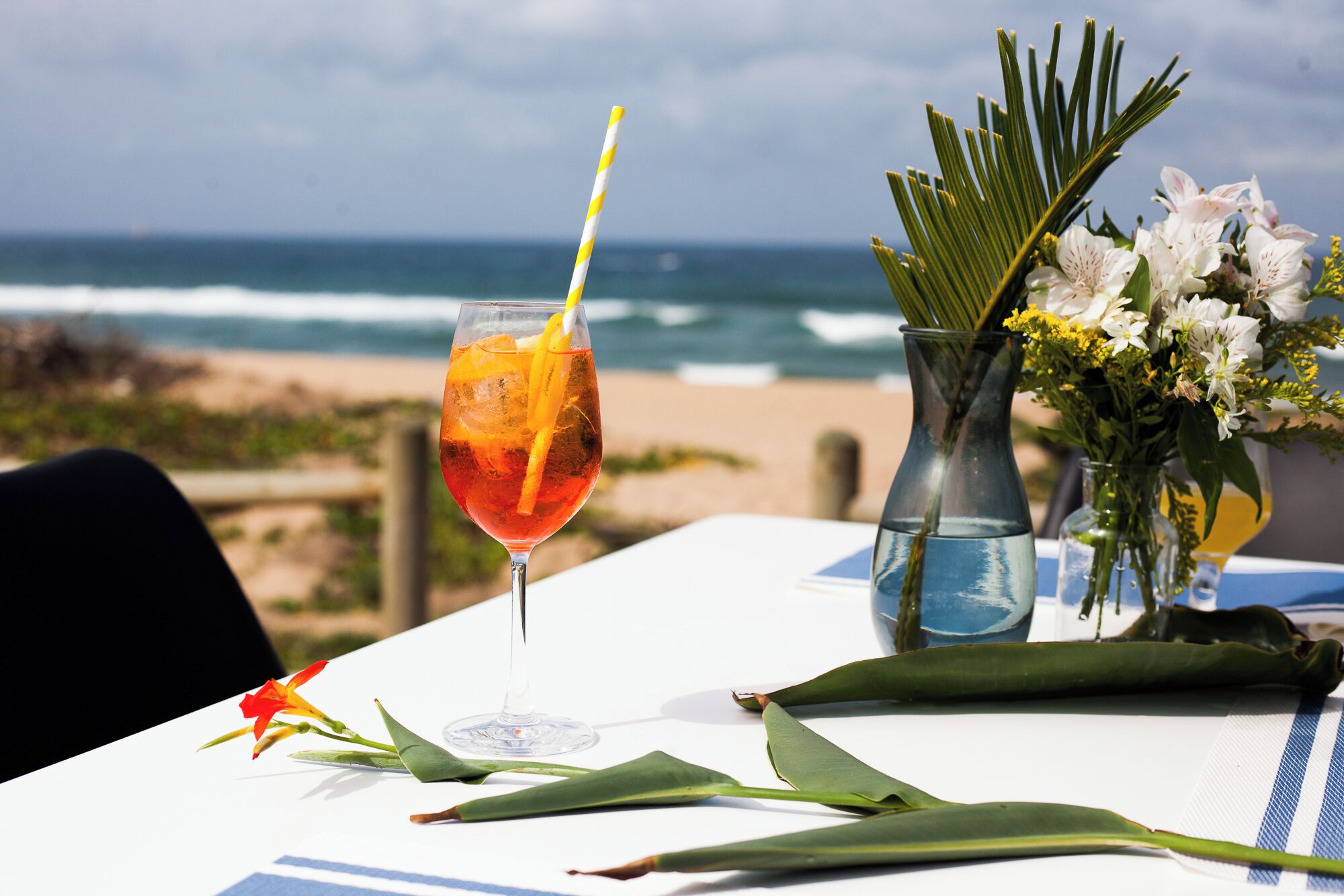 A table with a drink, a vase with flowers, and a view of the ocean.