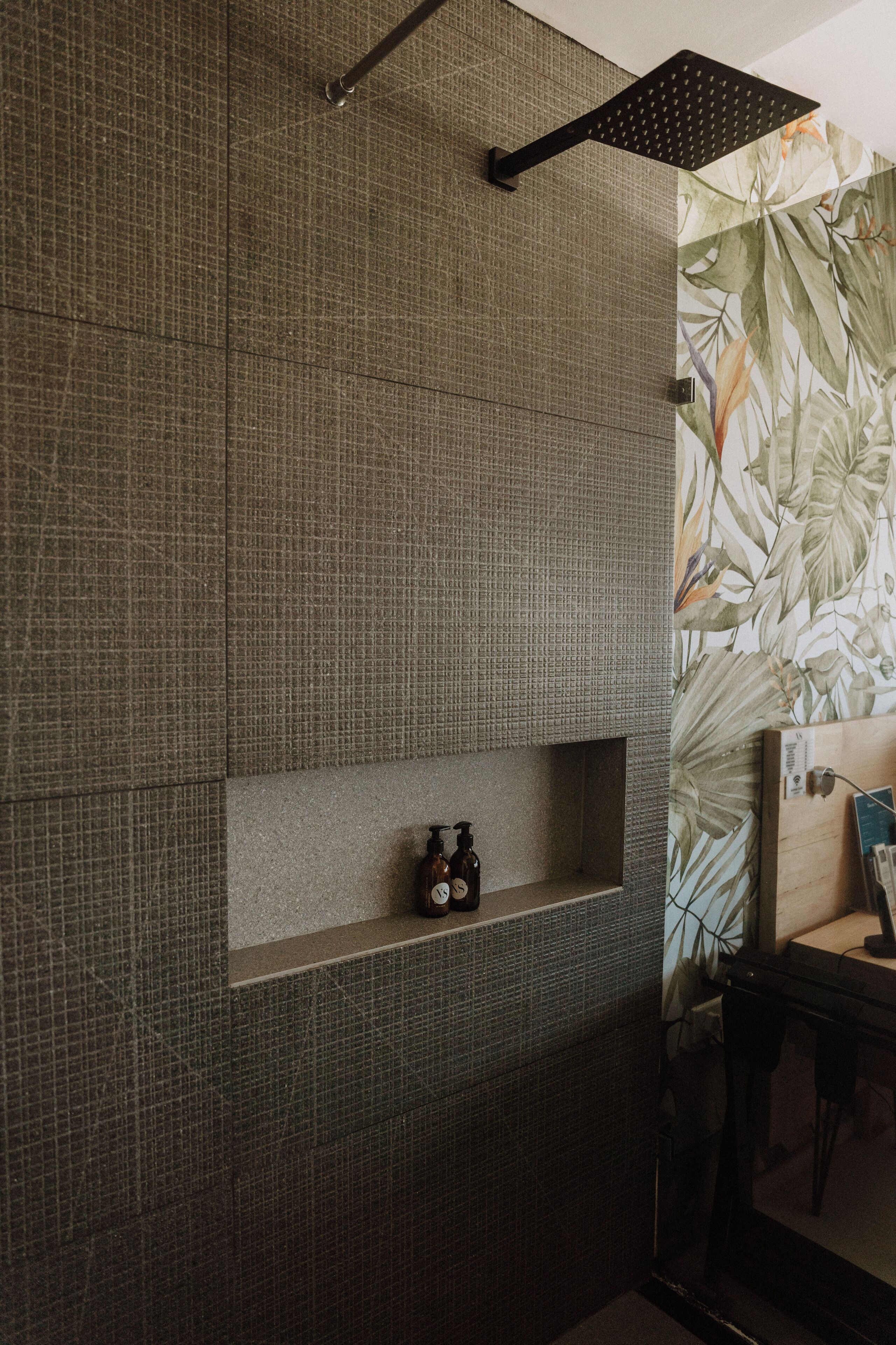 A bathroom with a textured wall, a shelf with a bottle, and a shower area with a floral wallpaper.