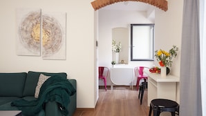 A living room with a green sofa, a coffee table, and a dining area with a white table and red chairs. There is a brick archway leading to the dining area, and a window with a view of the outside. - Casa Cosi - Maria Novella (Firenze)